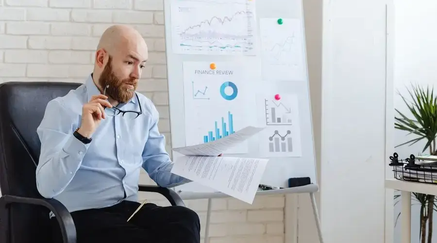 Bald businessman analyzing financial charts in modern office setting with whiteboard and paperwork.