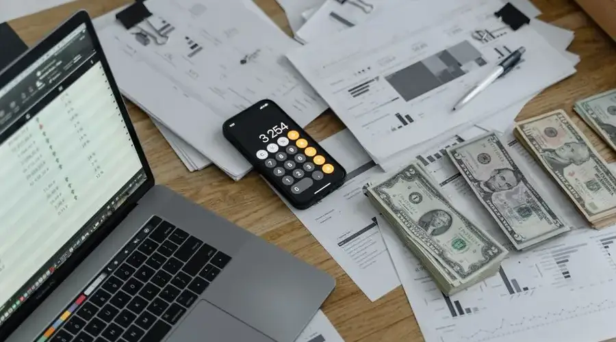 A desk with financial documents, currency, a laptop, and phone calculator.