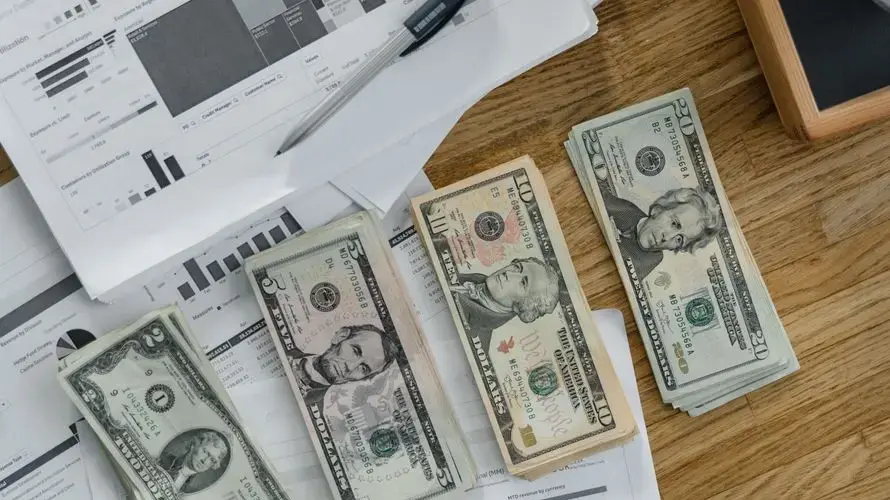 Stacks of US Dollar banknotes placed over financial documents and reports on a desk.
