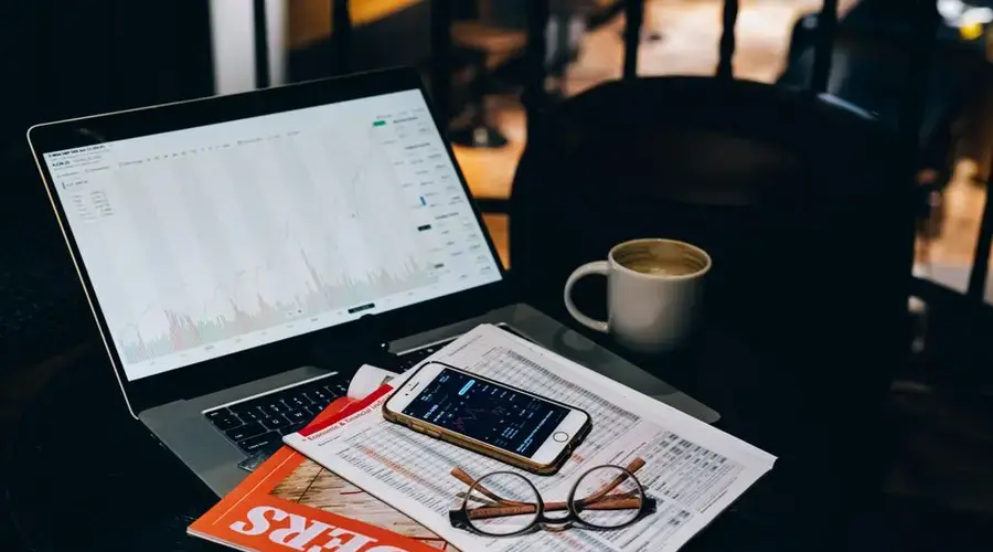A cozy office desk setup with a laptop displaying stock charts, coffee, and documents.