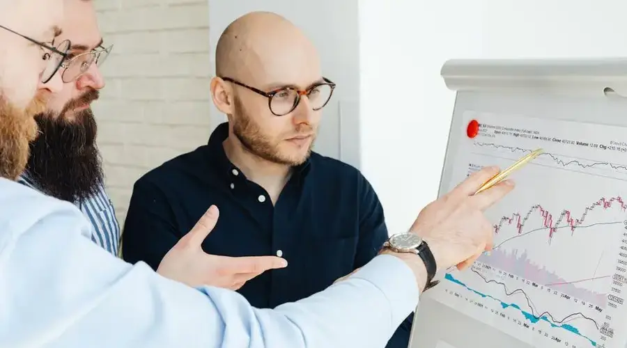 Three men discussing financial charts on a whiteboard during a business meeting.