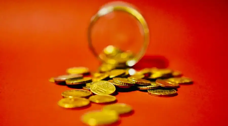 A collection of golden coins spilling from a glass jar on a vibrant red background symbolizes wealth.