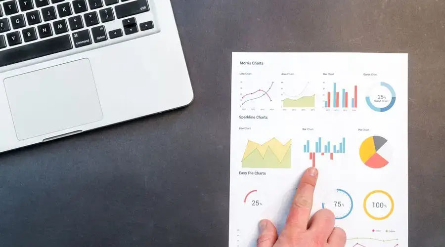 A hand pointing at colorful charts with a laptop in the background on a desk.