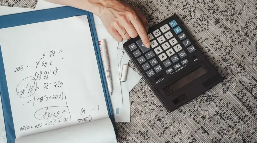 Top view of a hand calculating numbers with a large calculator and notes on a desk.