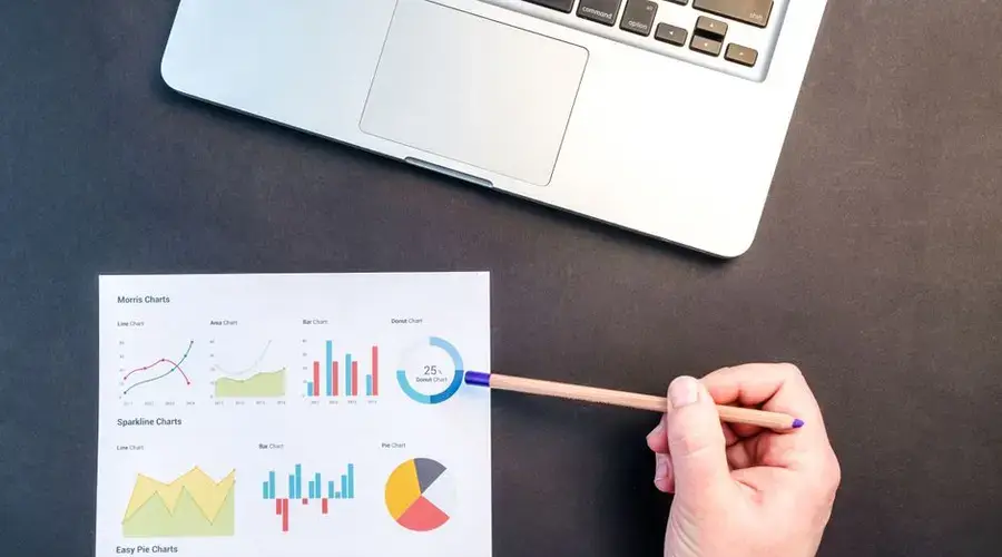 Hand holding pencil reviewing colorful data charts on desk with laptop.