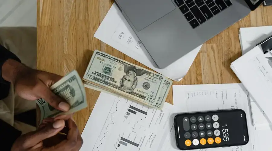 Close-up of hands counting cash on desk with calculator, charts, and laptop, illustrating financial management.