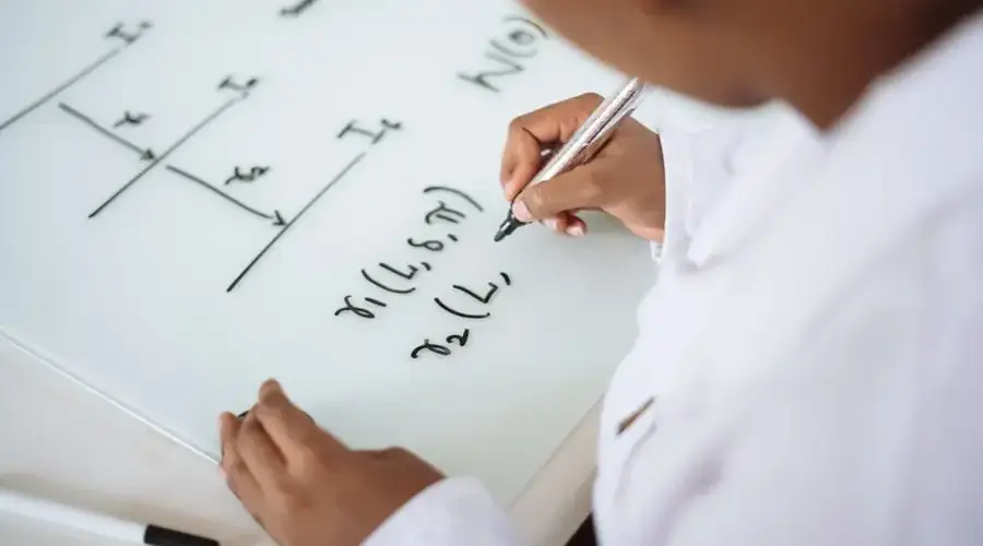 From above anonymous ethnic student wearing uniform and solving problem in chemistry while writing formula on white table in lab