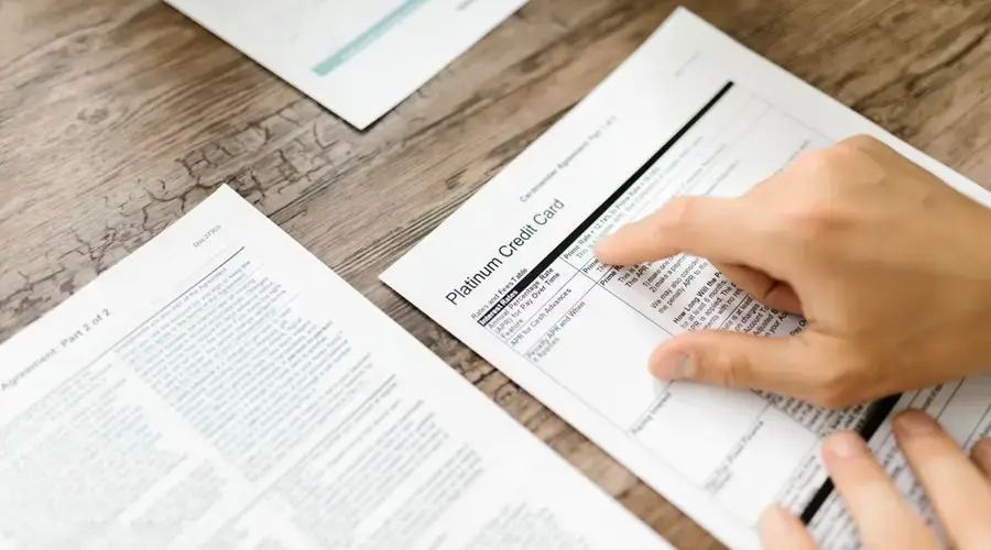 A hand examining a credit card agreement on a wooden desk, highlighting financial review.