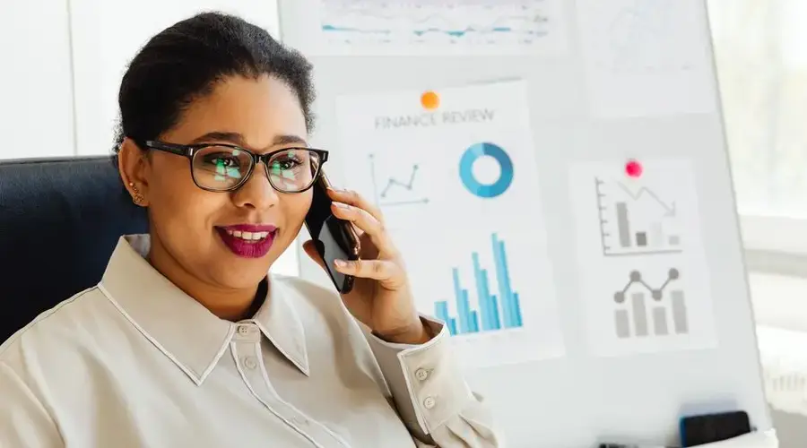 Confident businesswoman wearing glasses studies financial charts while talking on smartphone.