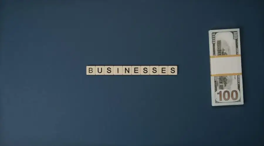 Dollar bills and 'BUSINESSES' spelled with tiles on a blue background, symbolizing finance.