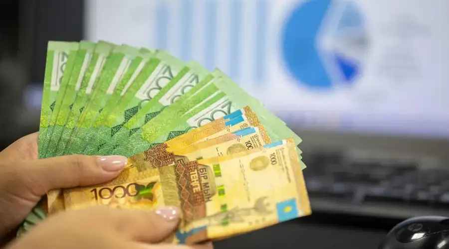 Close-up of hands holding Kazakhstani tenge bills with financial charts in background.