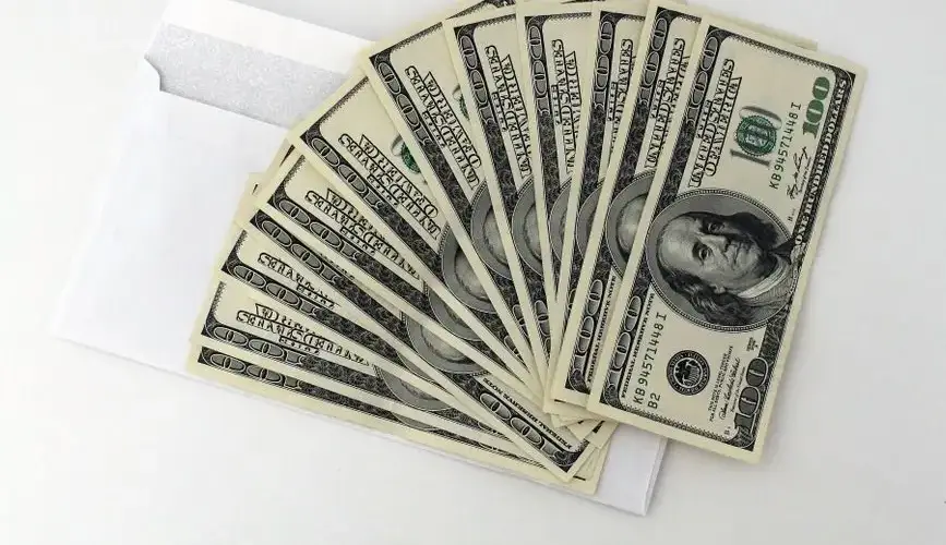 Fan of US $100 bills partially out of a white envelope on a white background.