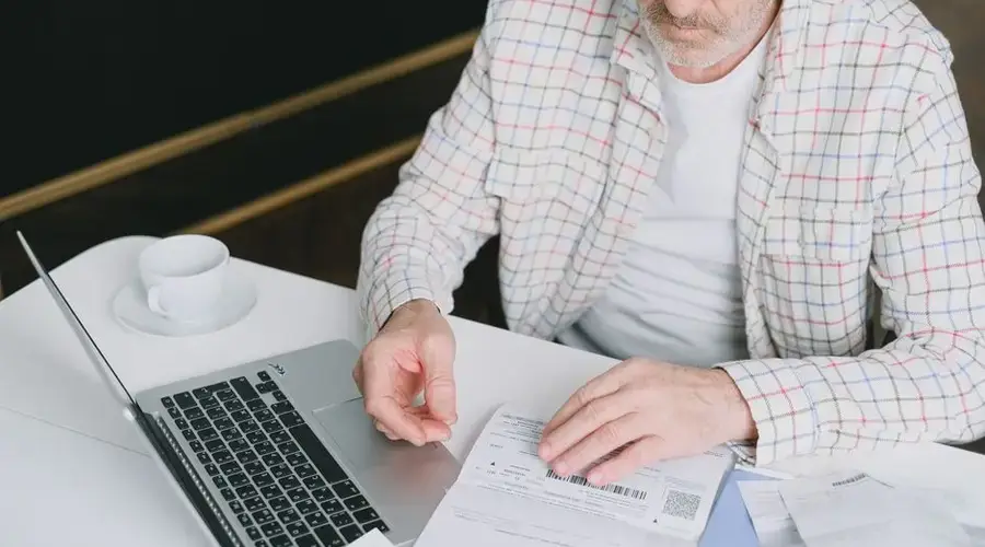 Elderly man with eyeglasses reviewing documents at a laptop. Indoor setting with natural light.