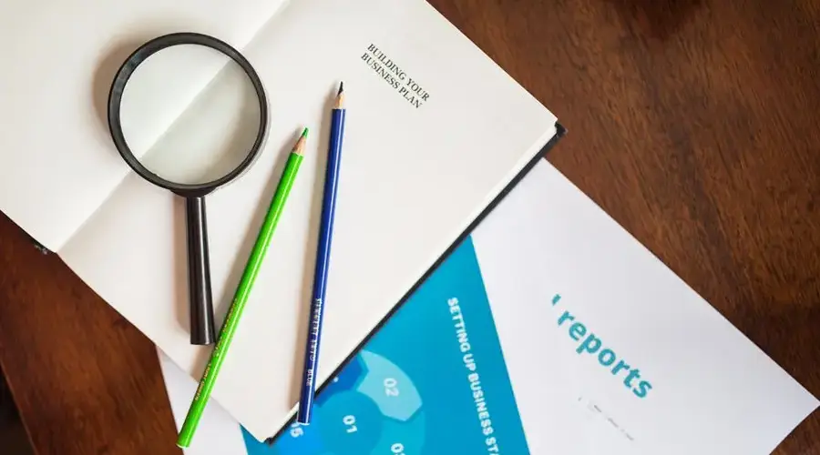 A top view of a magnifying glass, colored pencils, and business reports on a wooden table.
