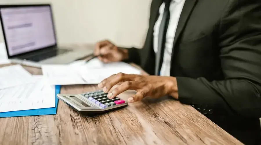 A businessman in a black suit using a calculator at a desk with financial documents and a laptop.