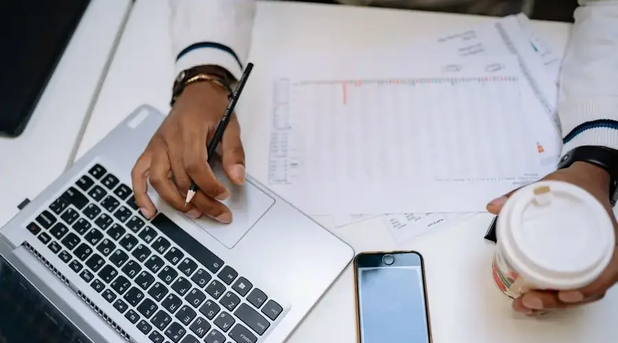 A businessman holding coffee and using a laptop at a desk with charts and a smartphone.