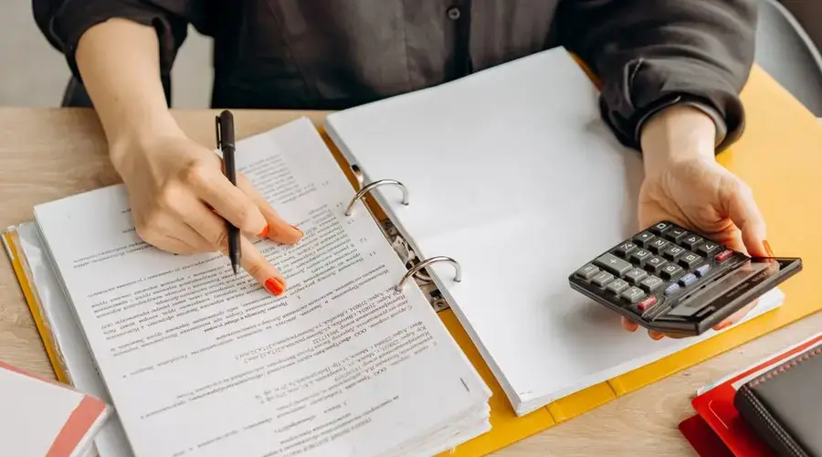 A woman reviewing documents and using a calculator on a wooden table.