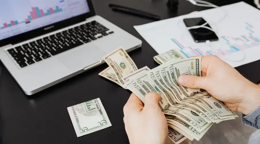 Person counting dollar bills at desk with laptop and financial charts, symbolizing finance management.