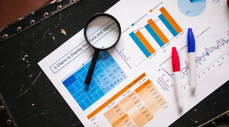 Flat lay of business charts and graphs with magnifying glass and markers on a dark surface.