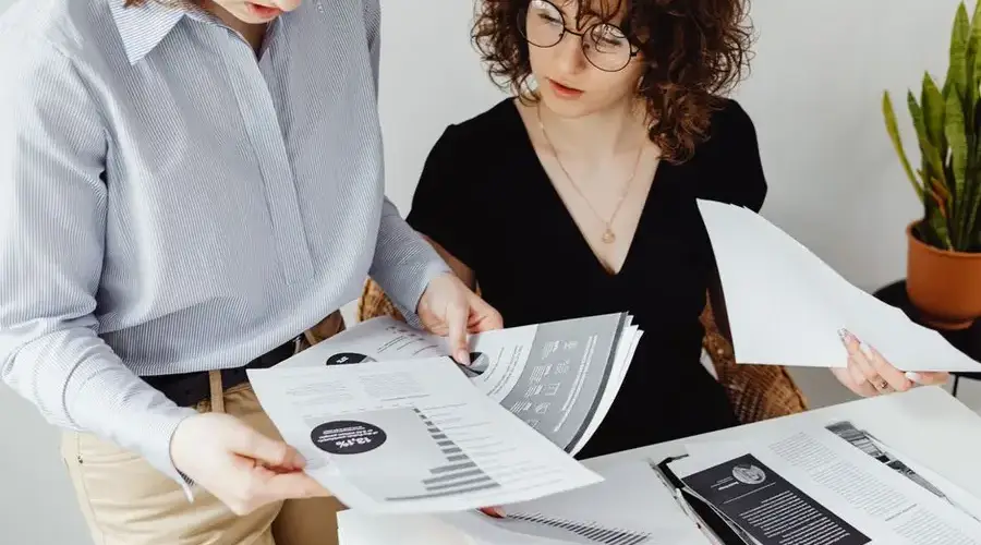Two women review and discuss financial documents in an office setting, highlighting teamwork and analysis.