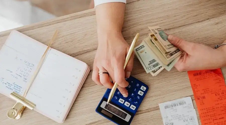 Hands managing finances with calculator, cash, and receipts on a wooden table. Ideal for budgeting concepts.