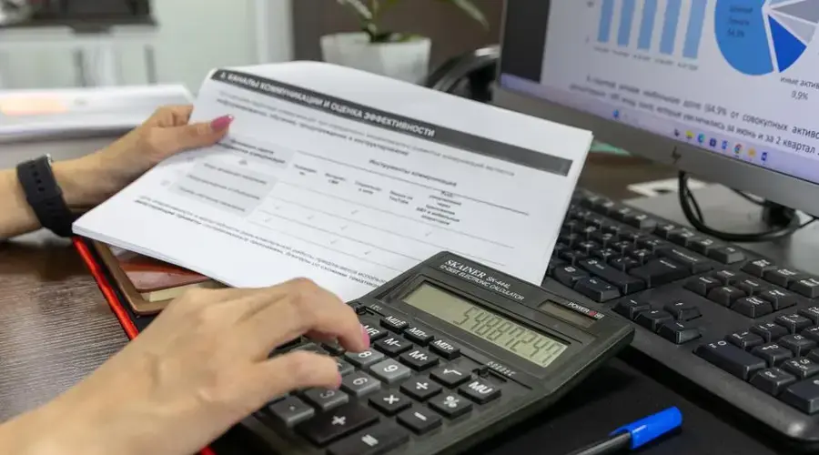 A person calculates financial data using a calculator and document, working at an office desk.