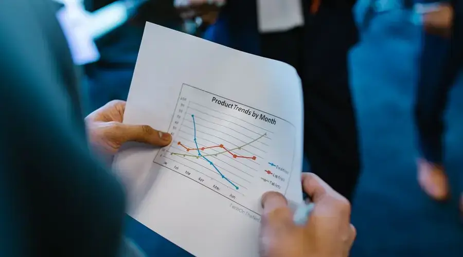 Close-up of hands holding a paper with a line graph showing product trends by month during a business meeting.