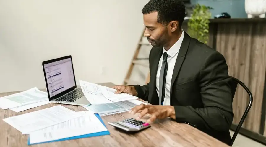 A businessman in a suit working on documents and using a laptop at an office desk.