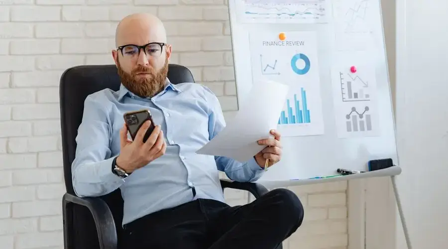 Bald businessman in corporate attire reviewing paperwork and smartphone data indoors.