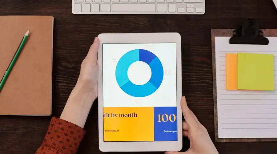 Tablet showing pie chart on desk with hands, keyboard, and notepad in office setting.