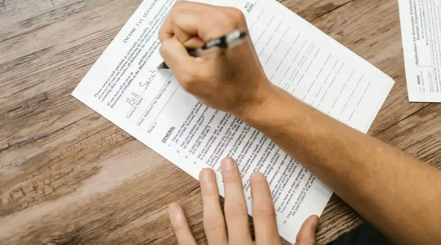 Person filling out a tax questionnaire with a pen, showing close-up on wooden table.