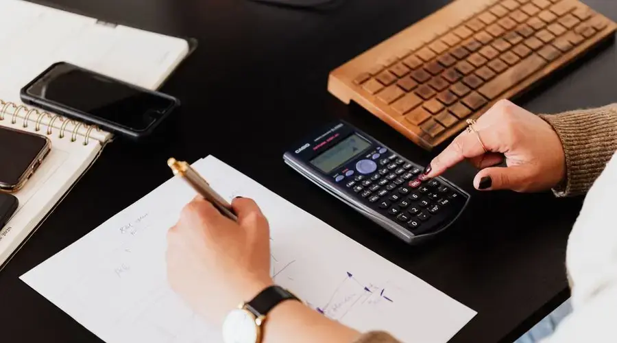 From above crop unrecognizable female accountant wearing stylish watches using calculator and writing down information on paper while working at desk with gadgets