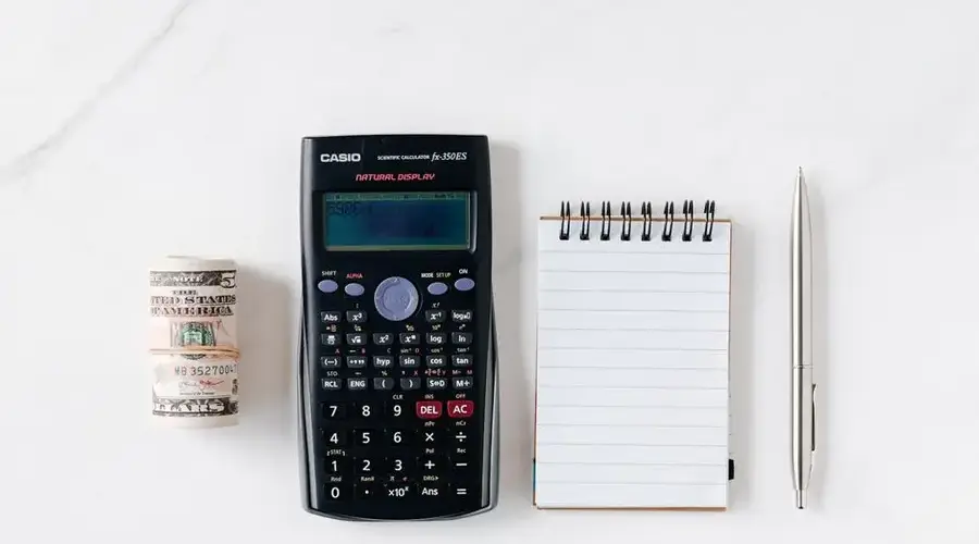 From above of roll of American dollar banknotes tied with rubber band near professional calculator and empty spring notepad with pen on marble table