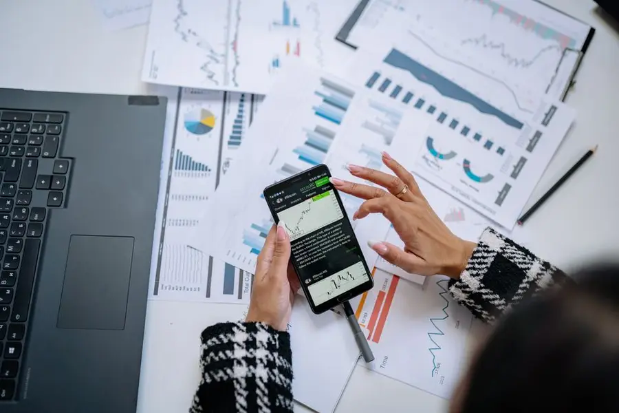 Businesswoman analyzing financial charts on smartphone with laptop and documents.