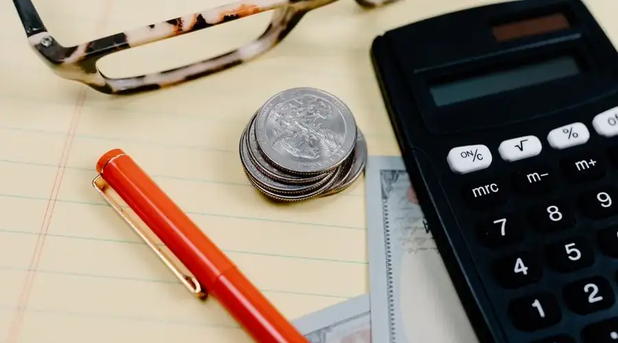 Close-up of a workspace with a calculator, coins, and glasses on a notepad.