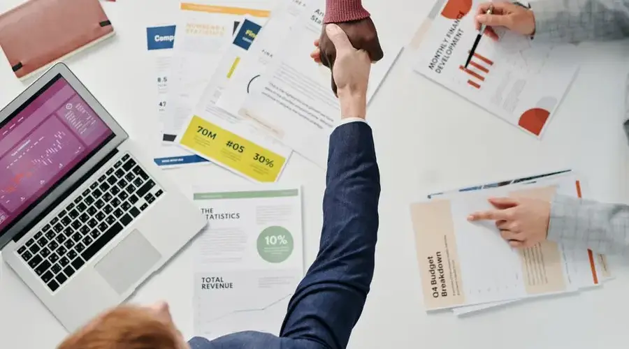 Business professionals engaging in a handshake over a desk filled with financial documents and a laptop.