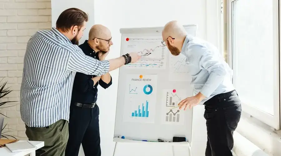 Three colleagues in a bright office discuss financial charts on a whiteboard.