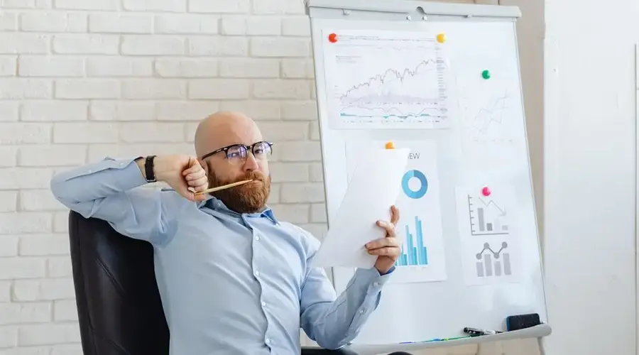 A businessman in glasses reviews financial charts, deep in thought by a whiteboard with graphs.
