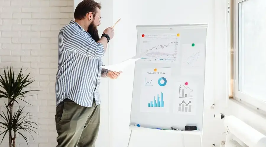 A man with a beard examines financial charts on a whiteboard in an office setting.