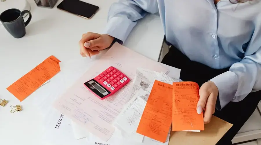 A woman reviews receipts and calculates expenses at a desk with a pink calculator.
