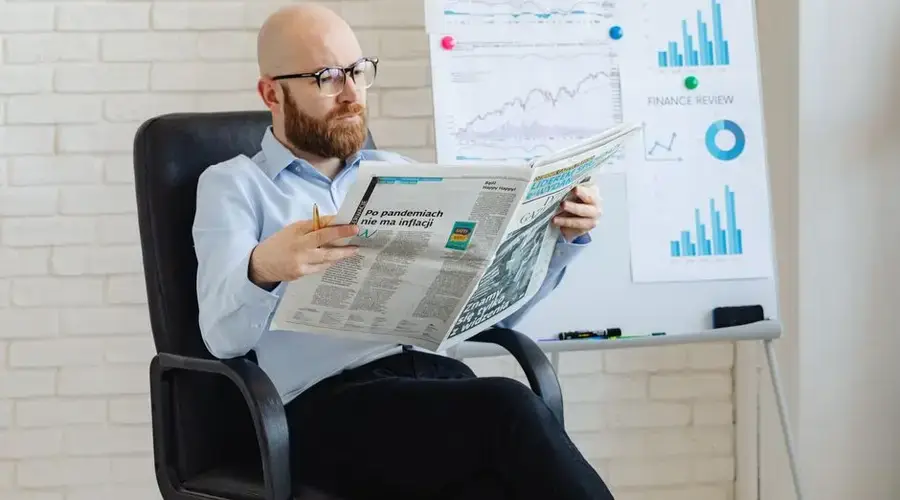 Bald man with beard reading newspaper in office with finance charts on whiteboard.