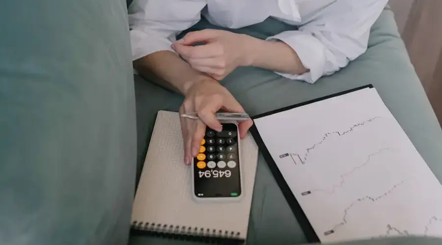A woman works indoors using a smartphone calculator and analyzing financial graphs, symbolizing modern finance.