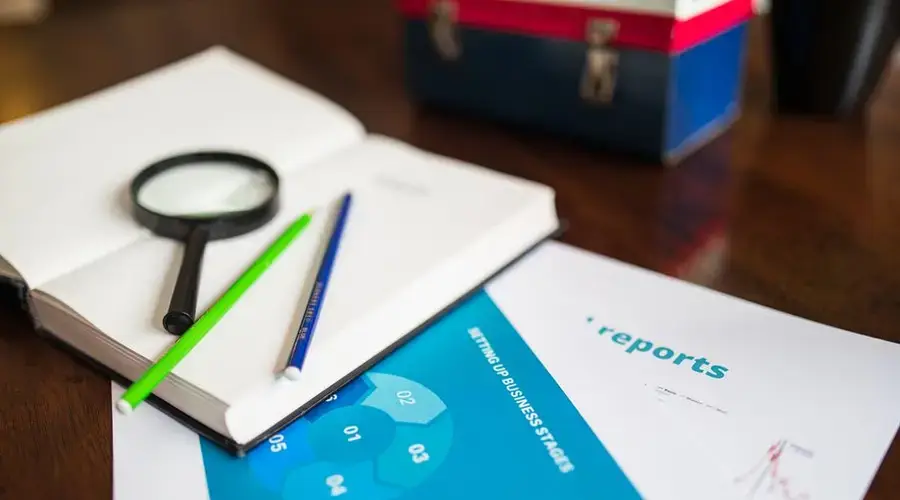 Open book with magnifying glass, pencils, and a business report on a wooden desk.
