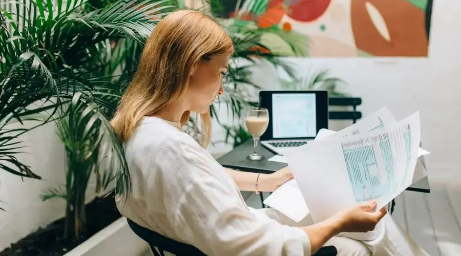 A woman reviews financial documents in an indoor garden space, with a laptop open.