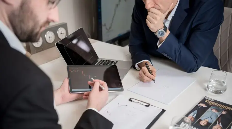 Two businessmen discussing financial charts on a tablet in a modern office setting.