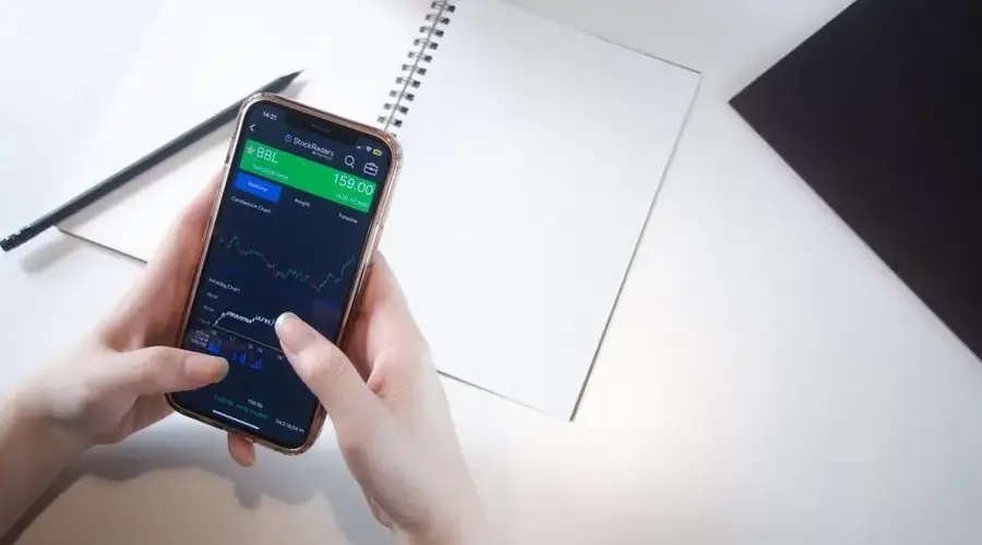 Close-up of hands using smartphone stock market app with notebook and pencil on desk.