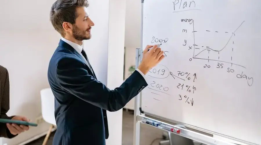 A businessman in a suit writes financial data on a whiteboard during an office planning session.