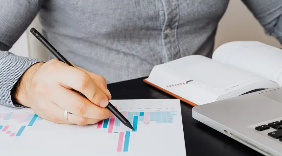 A business analyst examines a graph on paper with a laptop and notebook indoors.
