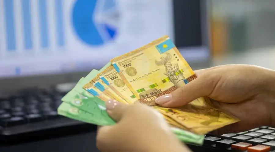 Human hands counting Kazakh tenge bills near a laptop displaying financial graphs.