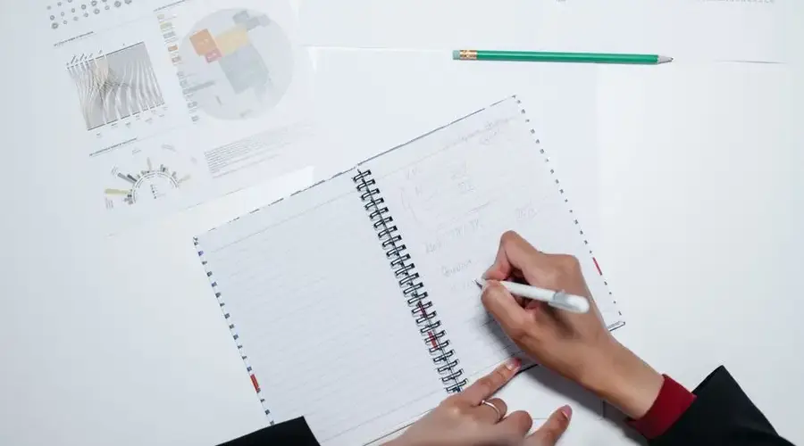 Hands writing in a notebook on a desk with charts, indicating office work.